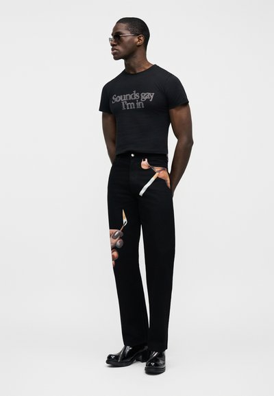 Μαύρο κοντομάνικο T-shirt με λευκή επιγραφή «Sounds gay I'm in», συνδυασμένο με μαύρα τζιν που έχουν τυπωμένο σχέδιο από χέρια που κρατούν ψαλίδια. Λαμπερά μαύρα παπούτσια.