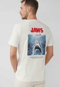 Biała bawełniana koszulka z dużym, kolorowym grafiką "Szczęki" na plecach, przedstawiająca rekina i pływaka z napisem poniżej.