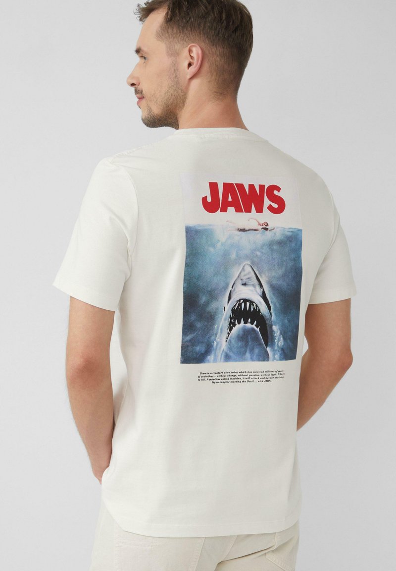 Biała bawełniana koszulka z dużym, kolorowym grafiką "Szczęki" na plecach, przedstawiająca rekina i pływaka z napisem poniżej.