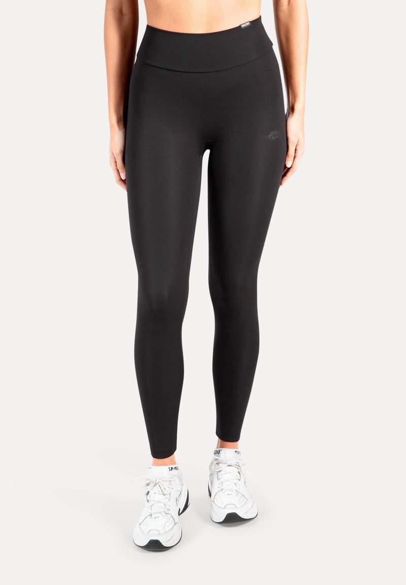 Zwarte legging met hoge taille en een gladde textuur, voorzien van een subtiel logo detail. Draagt witte sportschoenen.
