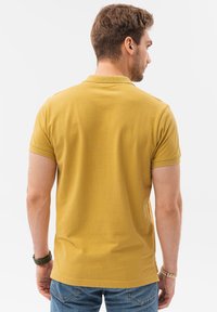 Polo giallo con maniche corte, dotato di colletto a costine e una trama liscia. Indossato con jeans blu, visto da dietro.