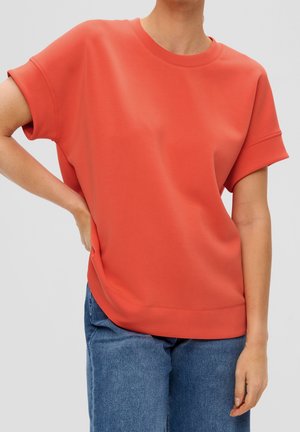 Osoba ubrana w luźną, krótką koralową bluzkę w połączeniu z niebieskimi dżinsami, z jedną ręką na biodrze i neutralnym jasnym tłem.