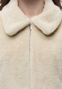Gros plan sur une personne portant une veste duveteuse couleur crème avec un grand col et une fermeture éclair argentée visible à l'avant.