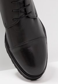 Scarpe da uomo in pelle verniciata nera con punta coperta e chiusura con lacci, viste da un'angolazione ravvicinata dall'alto su una superficie bianca.