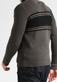 Mężczyzna w brązowym swetrze z teksturowanego dzianiny z czarnymi poziomymi paskami na górnej części pleców i ciemnych spodniach.