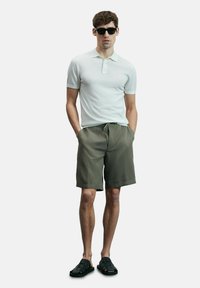 Witte poloshirt met korte mouwen, olijfgroene shorts met trekkoord en zwarte sandalen. Model staat met handen in de zakken tegen een effen achtergrond.