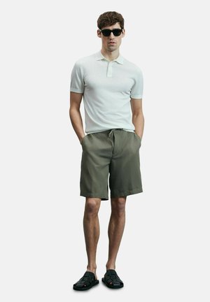 Witte poloshirt met korte mouwen, olijfgroene shorts met trekkoord en zwarte sandalen. Model staat met handen in de zakken tegen een effen achtergrond.
