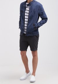 Marinblå lättviktsjacka med ståkrage och fickor med dragkedja, bärs över en vit och marinrandig skjorta, tillsammans med svarta shorts och vita skor.