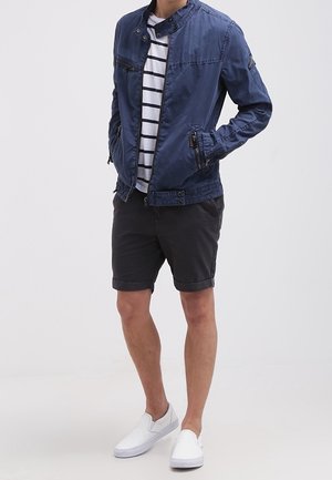 Lichtgewicht marineblau jasje met een staande kraag en ritszakken, gedragen over een wit en marineblauw gestreept shirt, gecombineerd met zwarte shorts en witte schoenen.