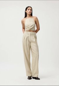Femeie îmbrăcată cu un top scurt halter bej și pantaloni largi cu talie înaltă, stând cu mâinile în spate pe un fundal simplu.