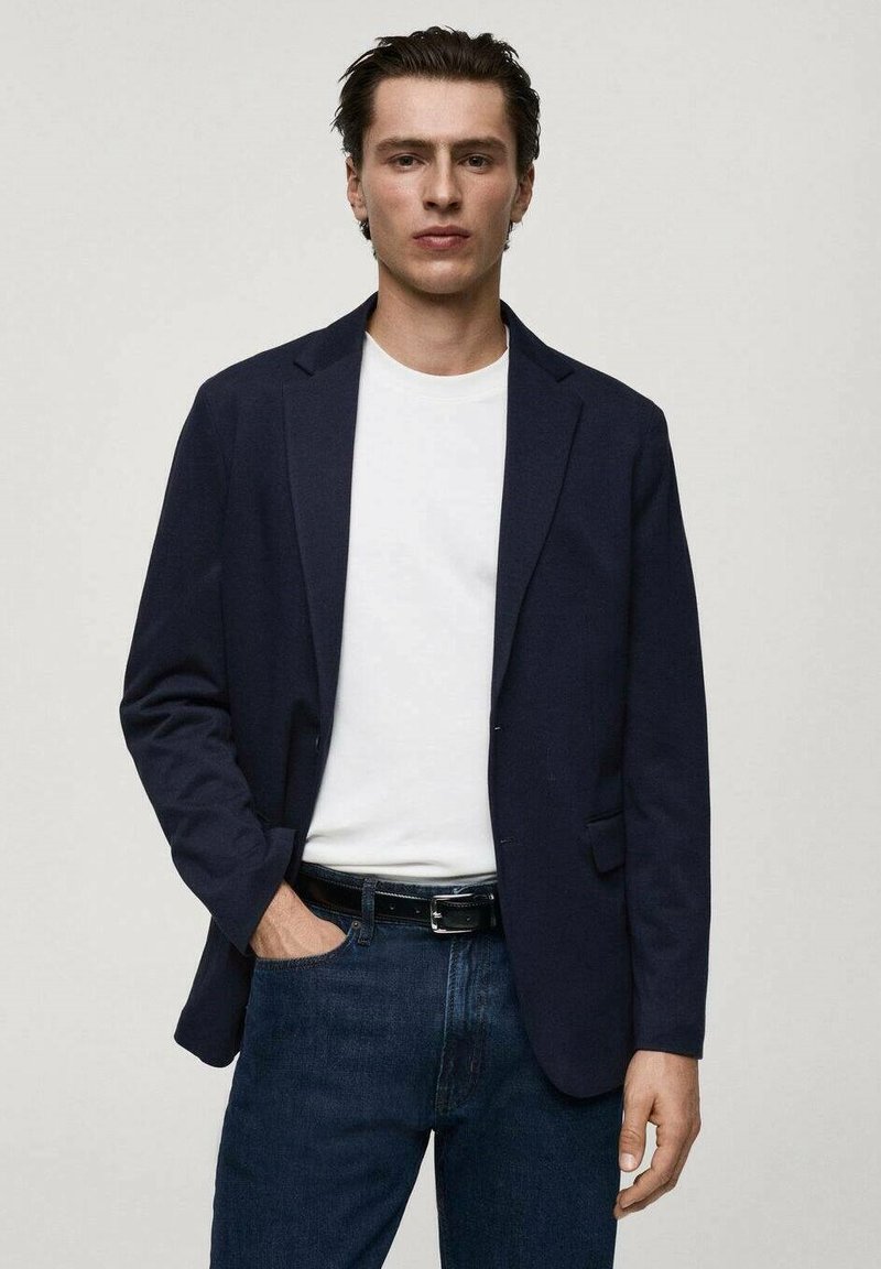 Dunkelblaue Blazer aus glattem Stoff, slim-fit Design, mit Reverskragen, kombiniert mit einem weißen T-Shirt und dunklen Jeans, steht in einer neutralen Umgebung.