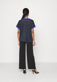 Granatowa koszulka o błyszczącej strukturze, z krótkimi rękawami i niebieskimi wykończeniami, zestawiona z czarnymi szerokimi spodniami i czarnymi sandałami na obcasie.