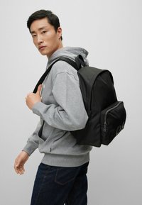 Sac à dos en tissu noir avec une forme structurée, poche avant zippée et grand logo en détail. Bretelles ajustables et finition texturée.