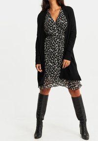 Svart lång kofta över en svart blommig klänning med korta ärmar. Knähöga svarta stövlar. Klänningen har en volangkant och en v-ringad hals.