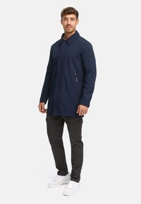 Marineblauer langer Mantel mit Kragen, Reißverschlusstaschen und strukturiertem Stoff. Kombiniert mit dunklen Hosen und weißen Sneakers.