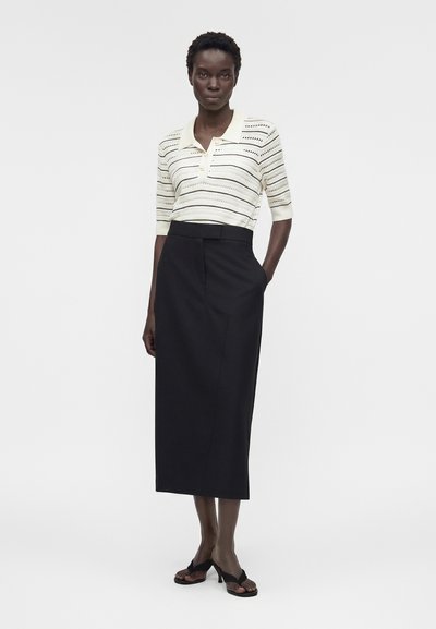 Γυναίκα που φοράει λευκή ριγέ πλεκτή πόλο με κοντά μανίκια και μαύρη midi φούστα με ψηλή μέση και μαύρα ανοιχτά τακούνια, στέκεται με τα χέρια στις τσέπες.