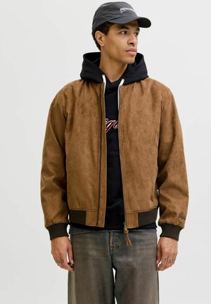 Jeune homme portant une veste en daim marron par-dessus un sweat à capuche noir, un jean gris et une casquette noire, regardant sur le côté devant un fond uni.