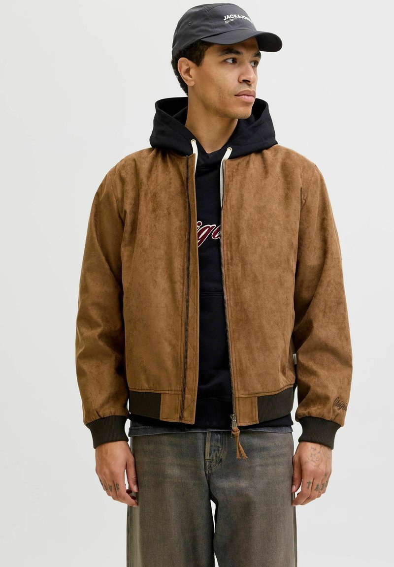 Jonge man die een bruine suède jas draagt over een zwarte hoodie, grijze spijkerbroek en een zwarte pet, kijkt opzij tegen een effen achtergrond.