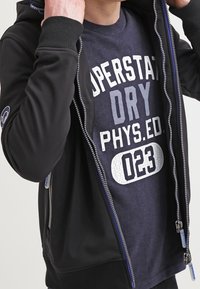 Svart zip-hoodie med texturerad yta, med en blå innerfoder och en grå grafisk tryckt t-shirt under. Ribbad mudd och fåll.