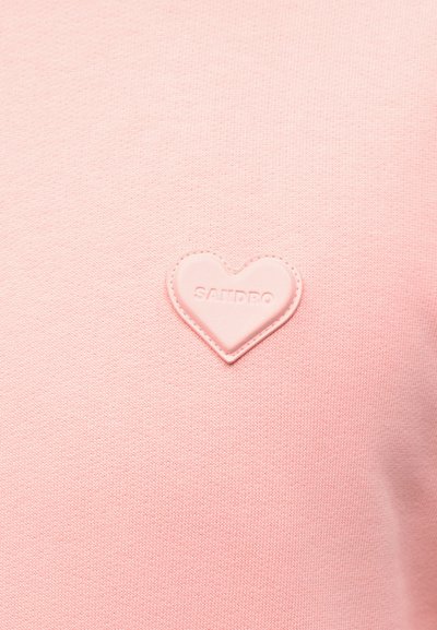 Ανοιχτό ροζ ύφασμα με ένα μικρό καρδιόσχημο κεντημένο πατς που εμφανίζει τη λέξη "SANDRO" στο κέντρο.