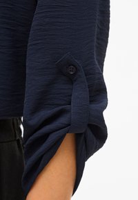 Μπλε ναυτικό πουκάμισο με υφή και μανίκια γυρισμένα προς τα πάνω, με κούμπωμα κουμπιού στο μανσέτο και απαλό, ελαφρύ ύφασμα.