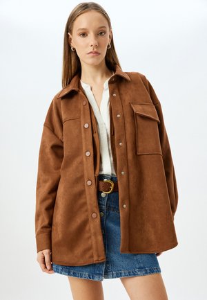 Brun ruskind skjortejakke med krave, trykknapper og en frontlomme, parret med en hvid top og en denimnederdel.