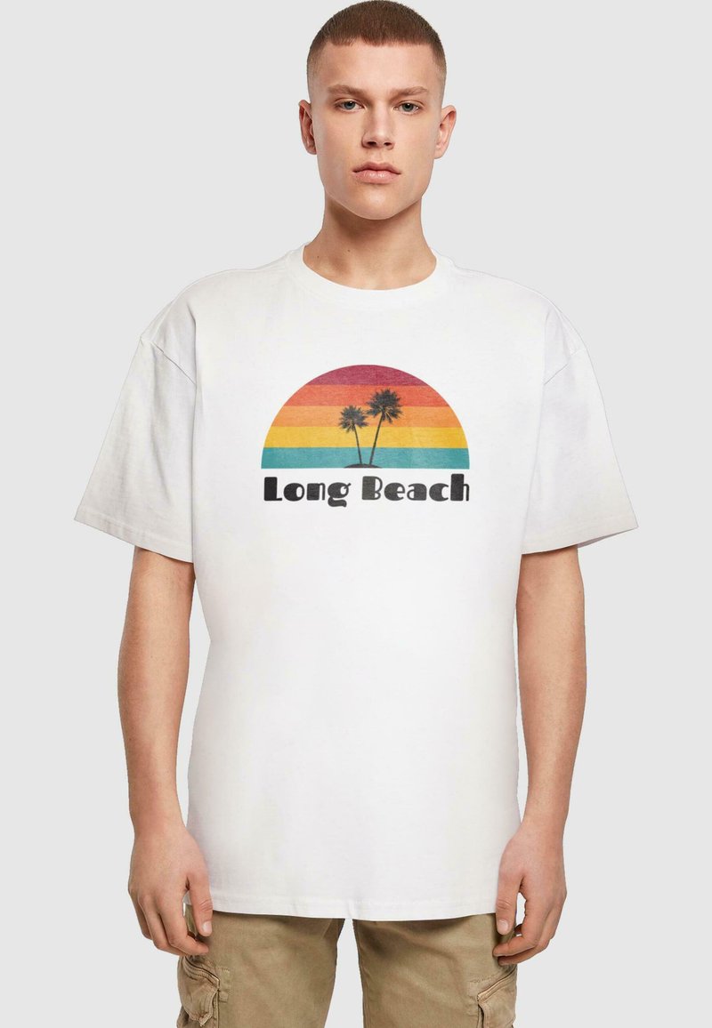 Valkoinen puuvillainen t-paita, jossa on värikäs grafiikka sateenkaaren auringonlaskusta ja palmuista, teksti "Long Beach" rohkealla mustalla fontilla.