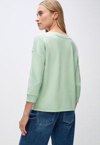 Lys grøn, langærmet top med ribbede detaljer på skuldrene, afslappet pasform, parret med blå jeans. Blød tekstur, enkelt design.