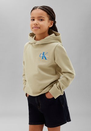 Junge Person mit geflochtenem Haar trägt einen beigen Calvin Klein Hoodie und schwarze Shorts, Hände in den Taschen, leicht lächelnd, steht vor grauem Hintergrund.