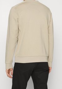 Felpa beige a maniche lunghe con scollo rotondo, caratterizzata da una texture liscia e polsini elasticizzati, indossata da una persona con pantaloni neri.
