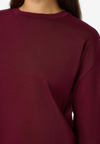 Camicetta bordeaux a maniche lunghe realizzata in tessuto morbido con scollo rotondo e vestibilità comoda, caratterizzata da cuciture minimum e senza motivi visibili.