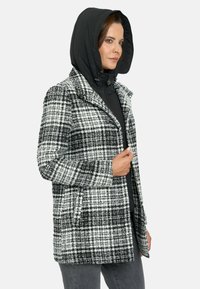 Abrigo de cuadros en blanco y negro con capucha negra desmontable; presenta grandes solapas y bolsillos laterales; tejido texturizado con un patrón prominente.