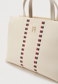 Μπεζ δερμάτινη τσάντα χειρός με δύο επάνω χεράκια, με κόκκινες και μαύρες βούλες κατά μήκος του κέντρου και με ένα χρυσό λογότυπο που προβάλλεται έντονα.