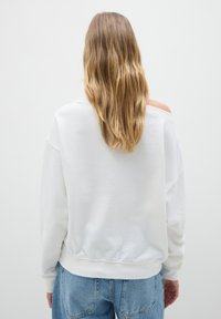 Persona con lunghi capelli biondi che indossa una felpa bianca a spalle scoperte e jeans blu, girata di spalle su uno sfondo bianco semplice.