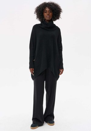 Czarny, oversized sweter z kapturem o luźnym fasonie i bocznymi kieszeniami, zestawiony z szerokimi czarnymi spodniami i ciemnym obuwiem.
