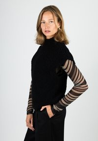 Black, textured sleeveless top with high neckline, paired with a sheer striped long-sleeve shirt. Model wears black pants.