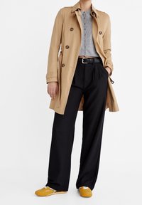 Trench beige con cierre de botones, suéter gris corto, pantalones negros de pierna ancha y zapatos amarillo mostaza con detalles en blanco.