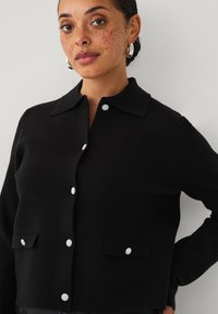 Zwarte gebreide cardigan met een kraaghalslijn, voorzien van vier decoratieve zilveren knopen en twee voorzakken met bijpassende accenten.