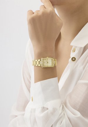 Poignet portant une montre rectangulaire en or avec des accents de diamants, associée à une chemise blanche à manches longues avec des boutons dorés, main reposant près du menton.