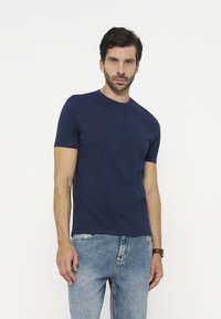 Uomo con capelli scuri e barba che indossa una maglietta blu navy a maniche corte semplice e jeans azzurri, in piedi davanti a uno sfondo bianco.