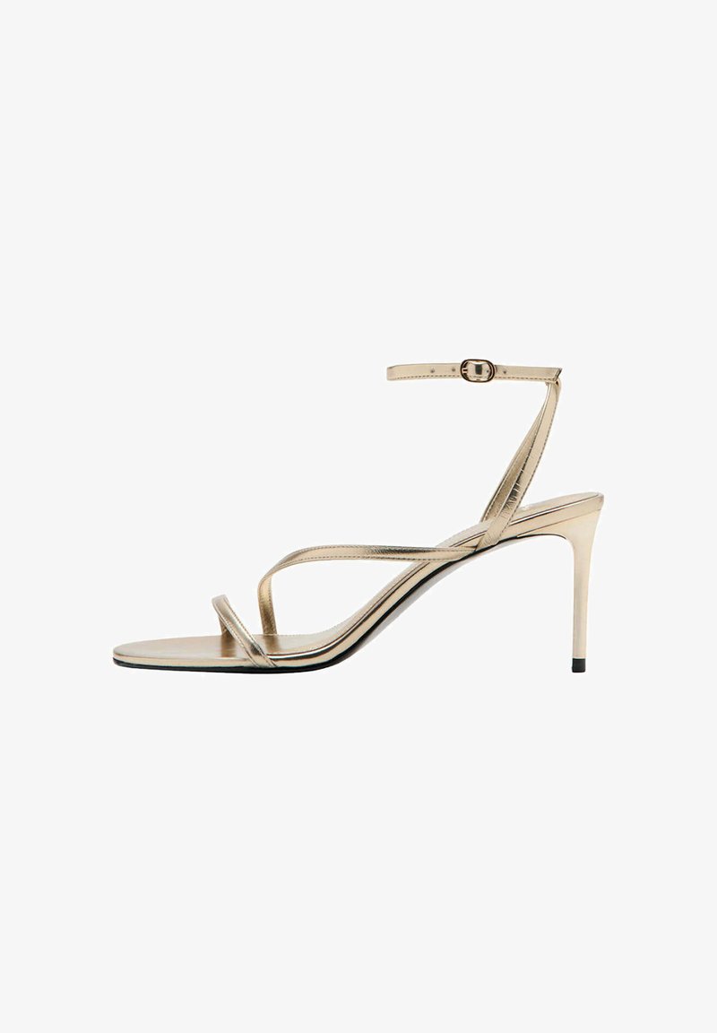 Sandale à talon haut dorée avec fines brides, bride de cheville fine avec boucle, talon fin et bout ouvert sur fond blanc.