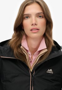 Zwarte waterdichte jas met capuchon, voorzien van oranje ritssluitingen en een Superdry-logo. Lichtroze shirt met een getextureerde kraag eronder.