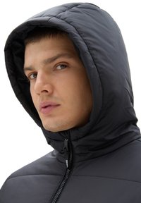 Czarna ocieplana kurtka z wysokim kołnierzem i kapturem. Posiada zamek błyskawiczny z przodu oraz pikowaną fakturę. Zbliżenie na twarz modelki.