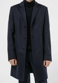 Abrigo de lana azul marino con cuello de muesca, cierre de botones sencillo, bolsillos laterales y un ajuste entallado. Cuello alto visible debajo.