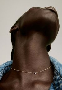 Collar de cadena de oro con un pequeño acento de piedra preciosa transparente, llevado alrededor del cuello contra piel oscura y tejido de mezclilla.