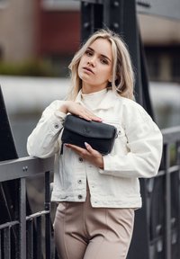 Černá kožená peněženka se zipovým uzávěrem, vybavená klopou a embosovaným logem, držena proti bílé džínové bundě a béžovým kalhotám.