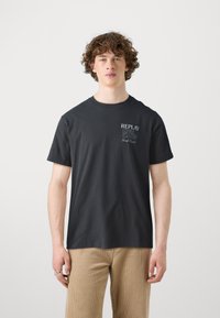 Svart bomullströja med rund halsringning och korta ärmar, med en tryckt grafik på vänster bröst som lyder "REPLAY HIGH STAKES Good Luck!"