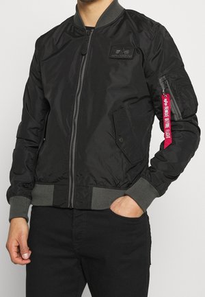 Mężczyzna noszący czarną kurtkę bombowiec Alpha Industries z szarymi prążkowanymi mankietami i pasem oraz czerwoną metką "Remove Before Flight" przy kieszeni na zamek na rękawie.