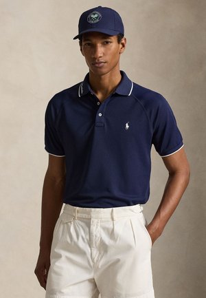 Mand iført navyblå polo-shirt med lille logo, hvide shorts og blå Wimbledon-kasket, stående med den ene hånd i lommen.