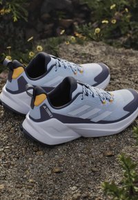 Ζευγάρι ανοιχτό μπλε και γκρι παπούτσια Adidas Terrex για τρέξιμο σε μονοπάτια με λευκές σόλες, που ξεκουράζονται σε χαλίκι με μικρά φυτά κοντά.